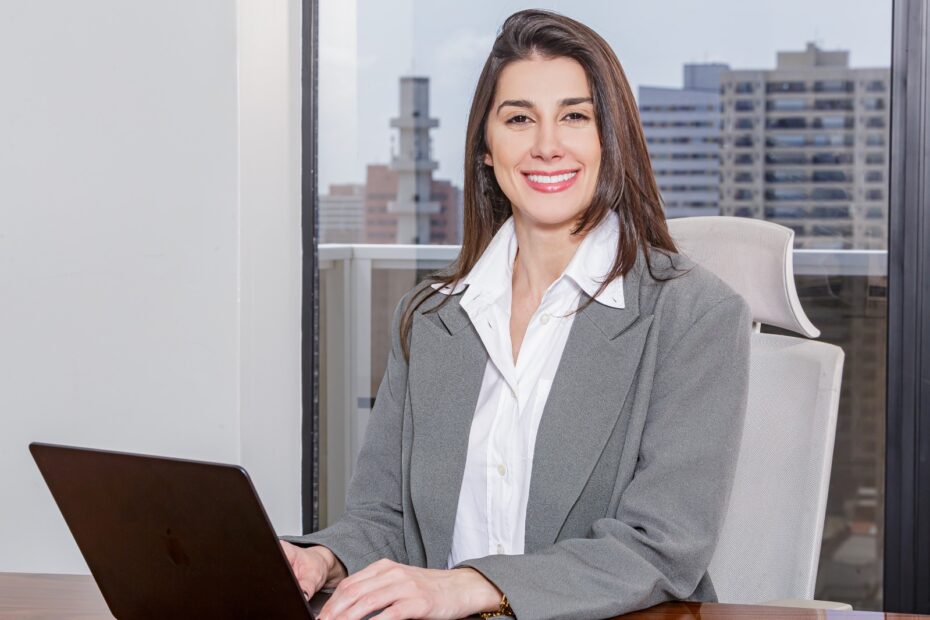 Foto de uma mulher com blazer social cinza e camisa branca sorrindo enquanto mexe no seu notbook